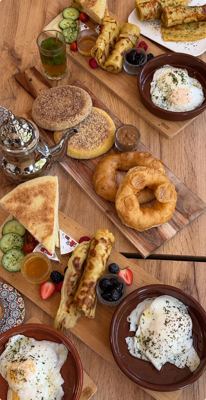 Marokkanische Frühstückstafel mit mehreren Holzbrettern: Spiegeleier, Msemen, Sfenj, Harcha, Oliven, Käse und Minztee